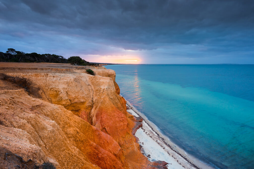 Large red cliffs overlooking a small beach and the sea, with the sun in the distance.
