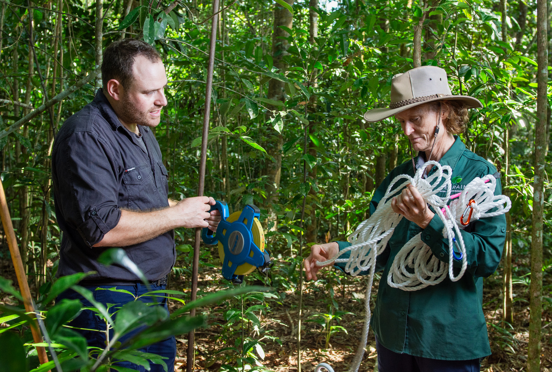 Two people standing in rainforest, one with rope in their hands the other with a large tape measure.