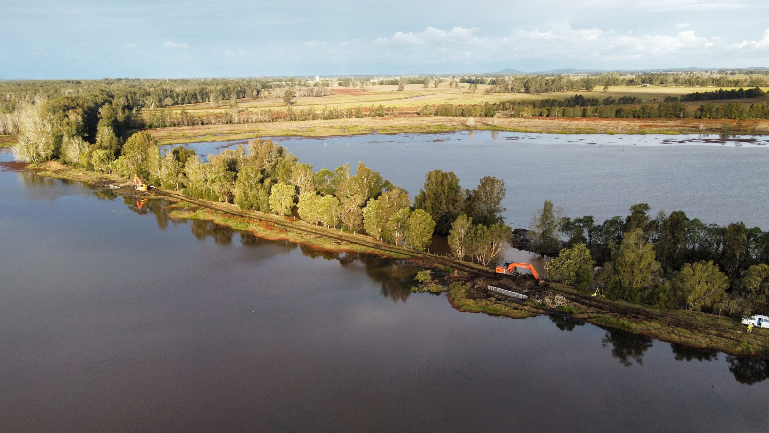 An aerial photo of a wetland with a spit of land running through it. Two excavators and a car can be seen clearing a road along the spit.
