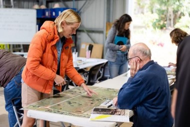 Woman standing at a table with a man sitting, looking at a map.
