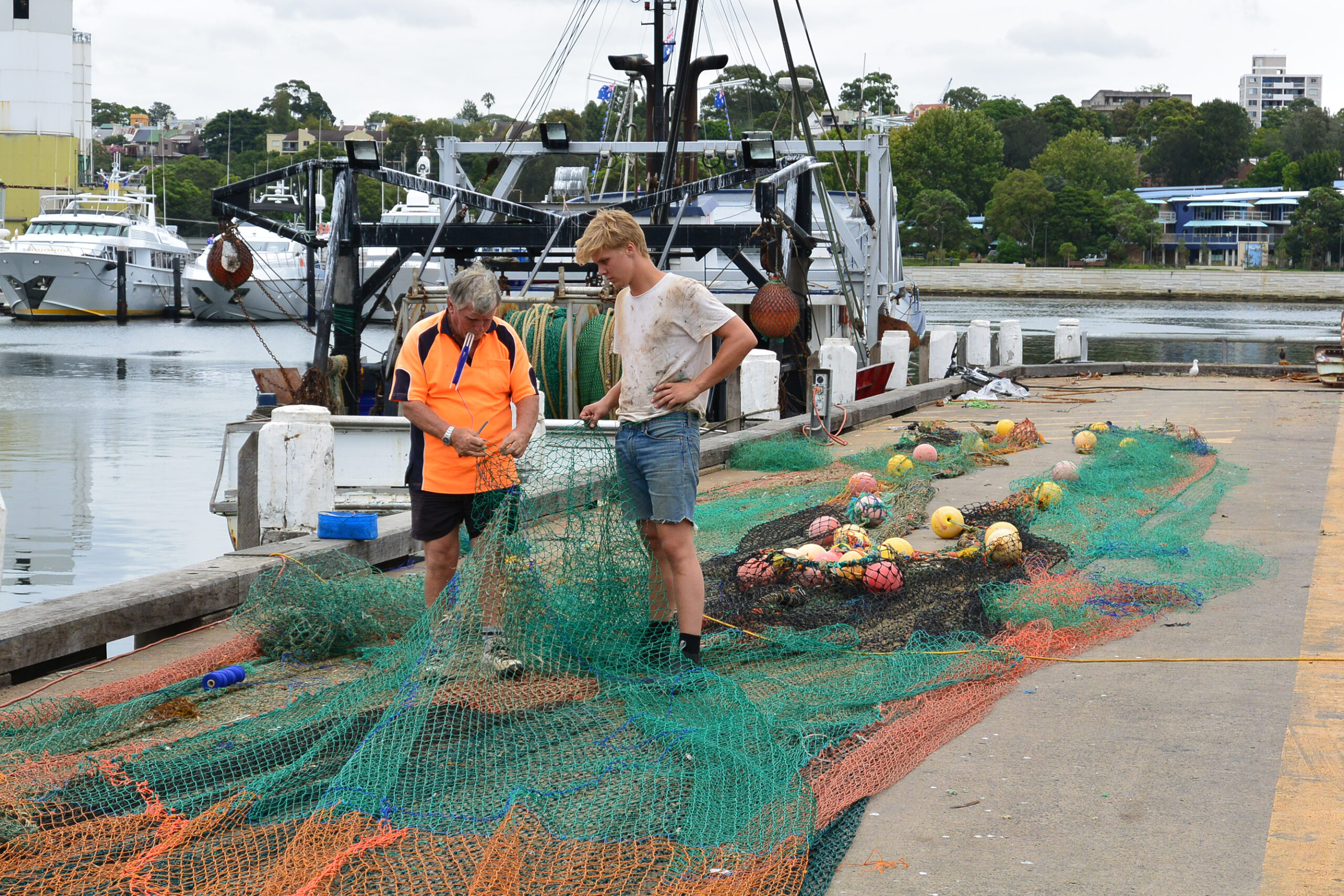 Two people standing in front of a moored boat, with nets stretched out along the length of the wharf.