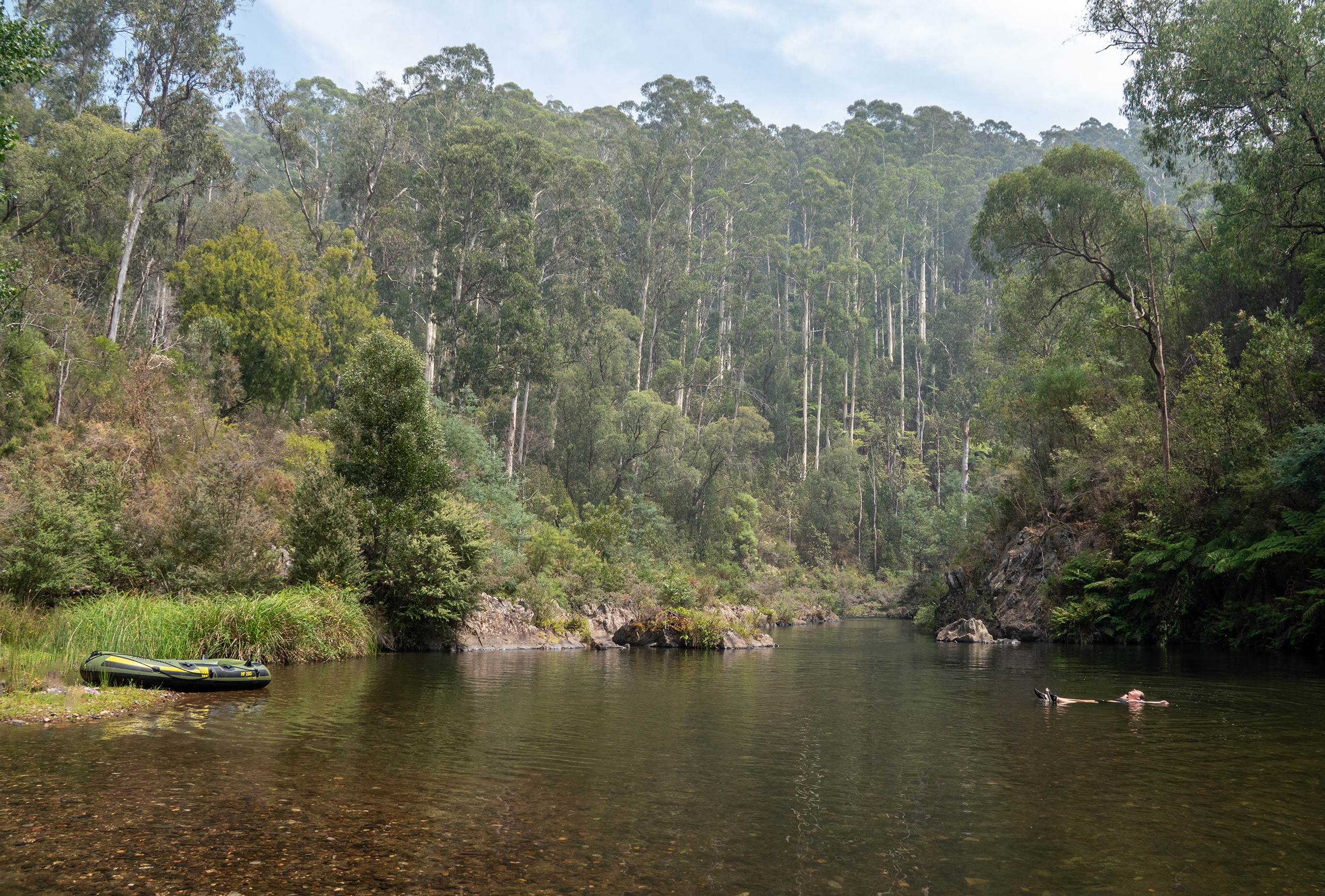 A river surrounded by tall trees, with a kayak on one bank and a man floating on his back in the centre of the water.