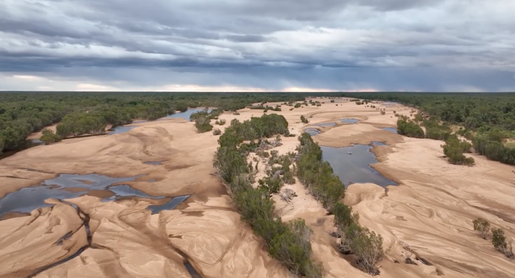 Long distance shot of open sand streaked with water runnels, vegetation lining the river bed and storm clouds overhead.