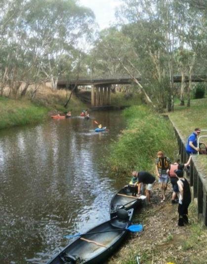 A group of people on a riverbank with two kayaks pulled up beside them. In the distance three more kayaks are out on the water, near a large bridge.