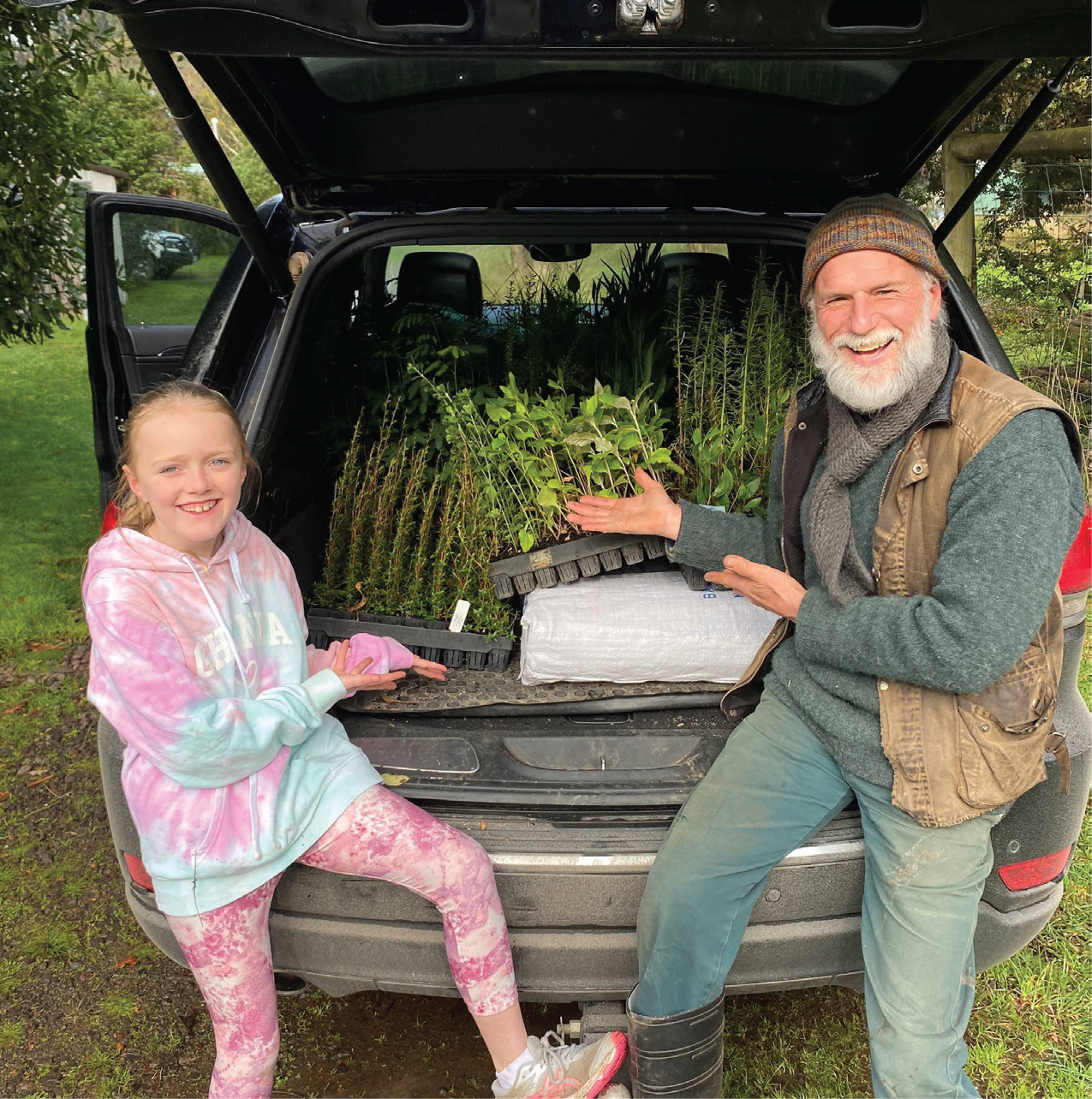 Two people sitting in the boot of a car, with a load of small saplings behind them.