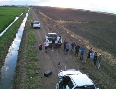 Group of people standing on a road between two paddocks; one with a rice crop and the other recently ploughed.