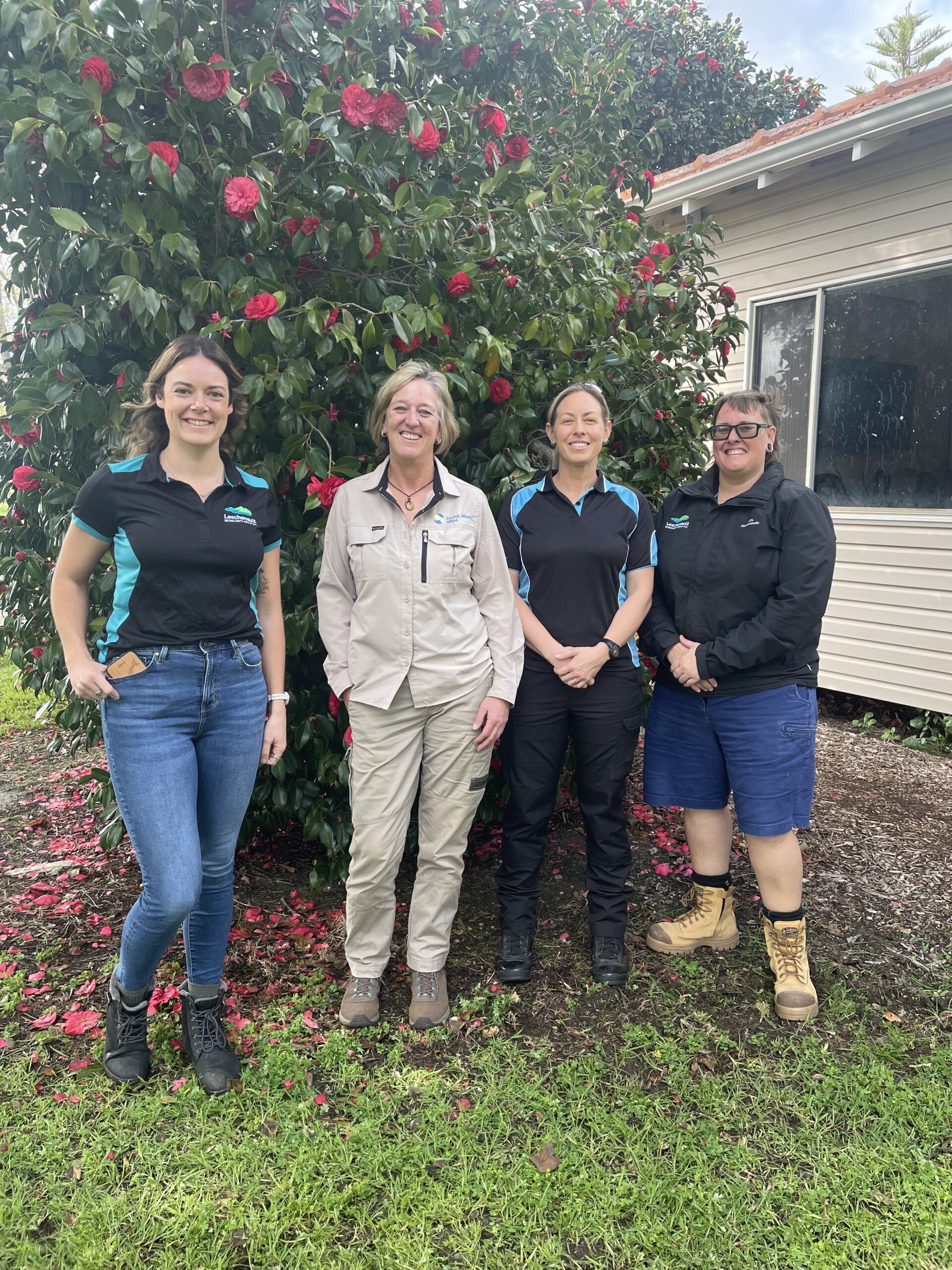 Four people are standing in front of a large bush, with red flowers, there is a cream-coloured building next to them.