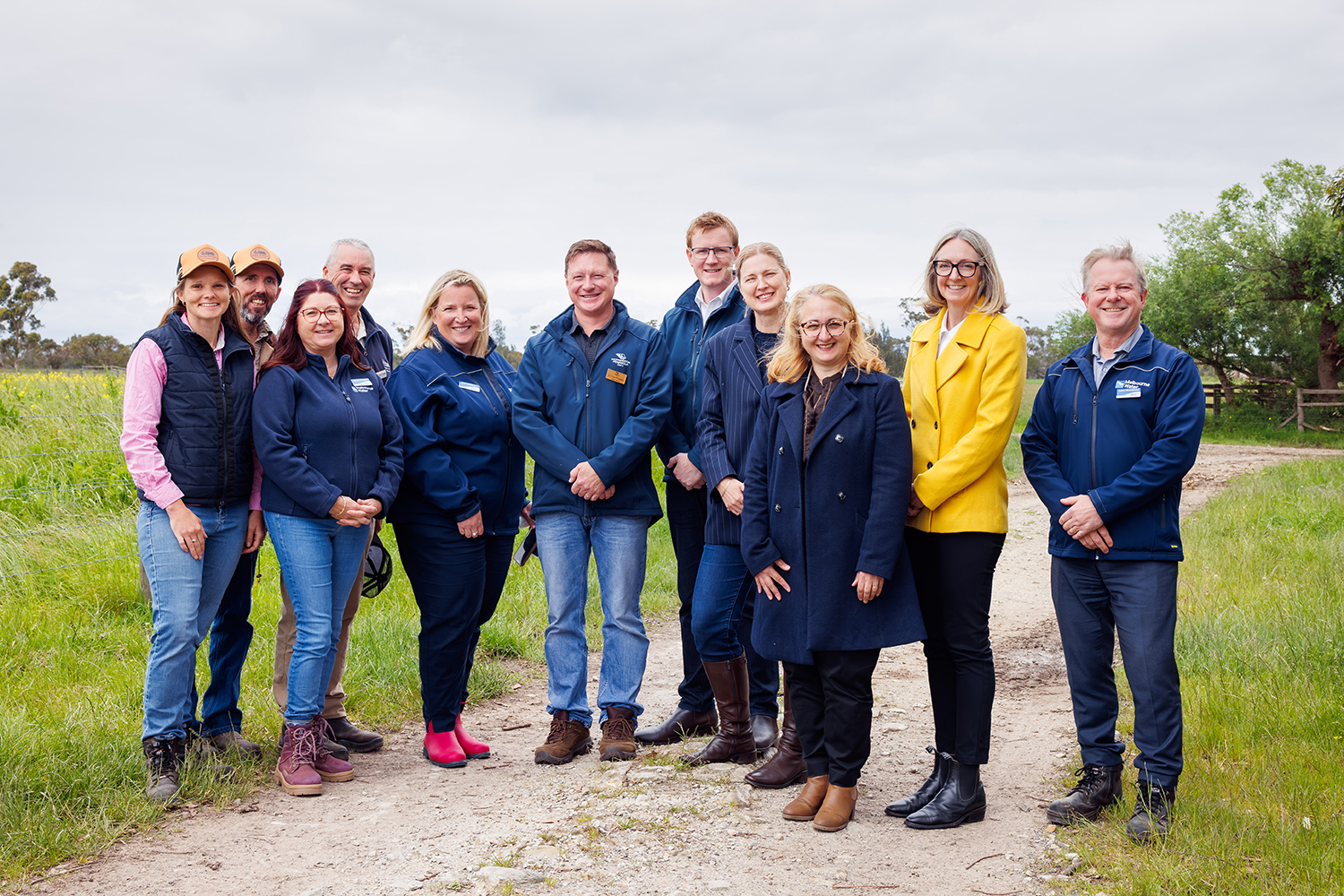 Group of eleven people looking at camera standing on path with grass either side and fence and trees in background.