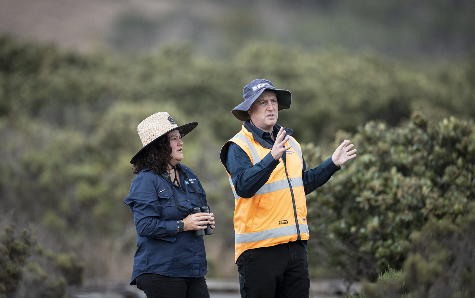 Two people standing with bushland around looking out of camera direction.