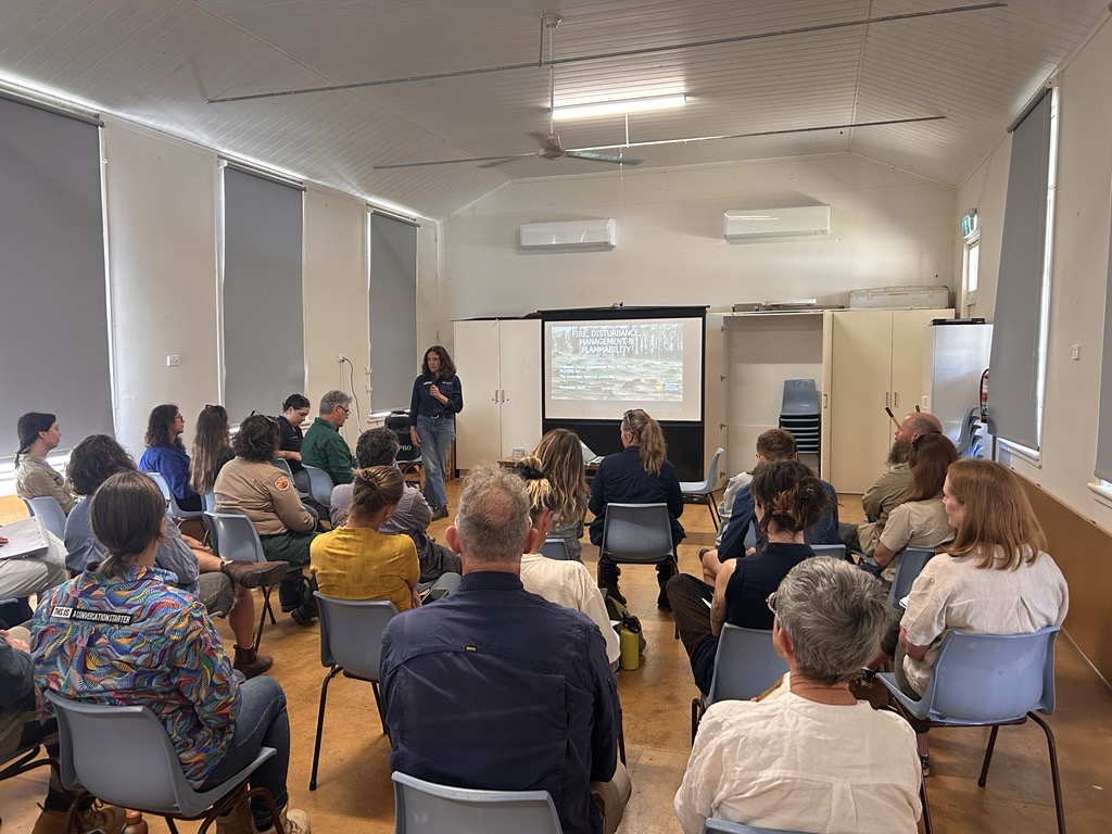 A group of people sitting on chairs, in a room watching a person present, with a screen behind them.