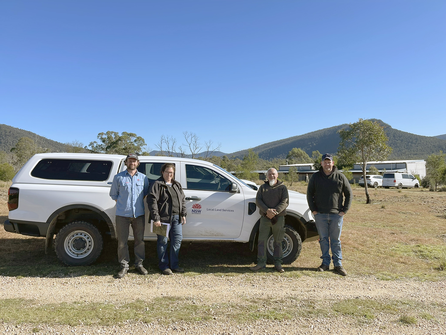 Four people standing in front of a car, with mountains and trees in the background.
