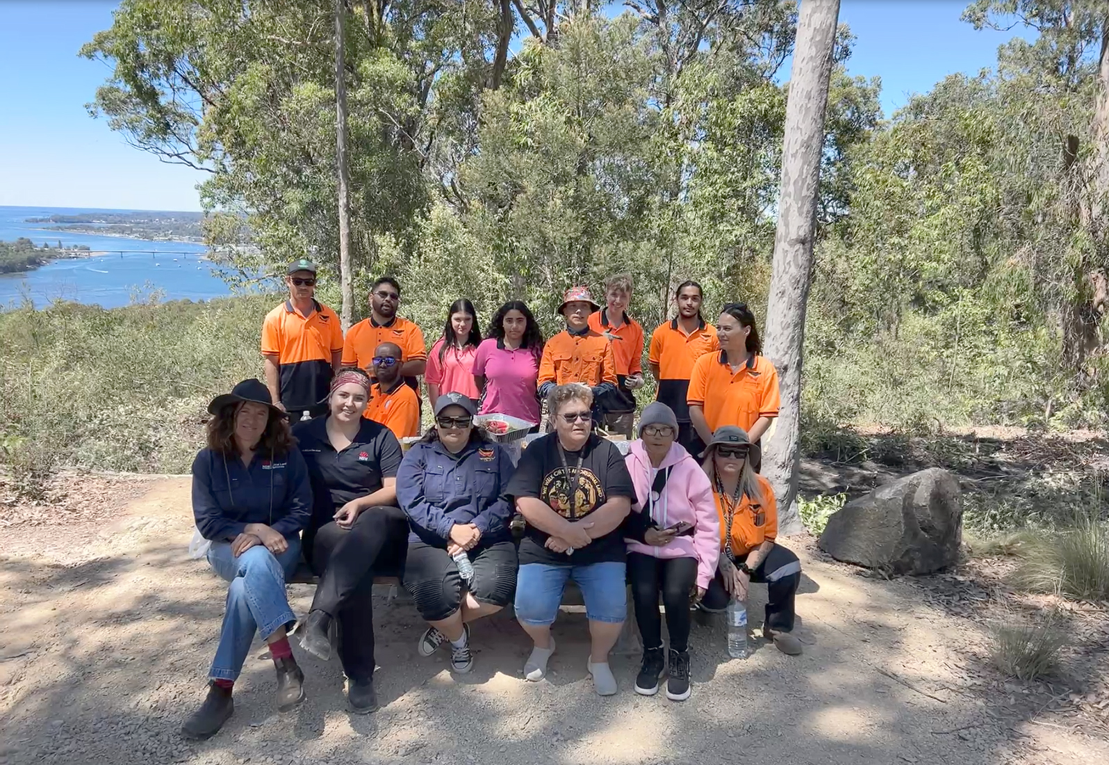 A group of people in high vis posing for a photo, in the background is bushland and the ocean.