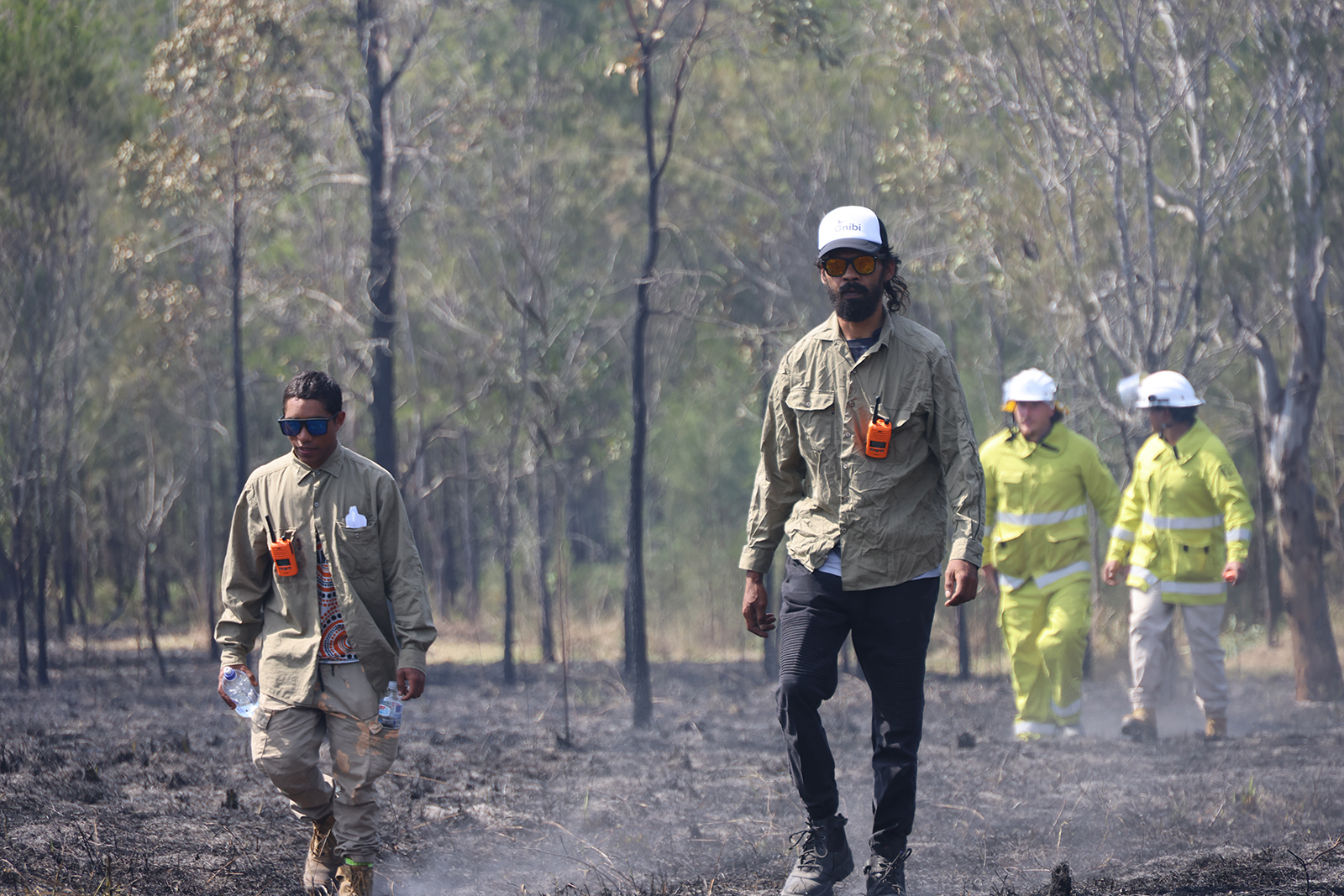 Four people walking on burnt bushlands ground, in a bush landscape.