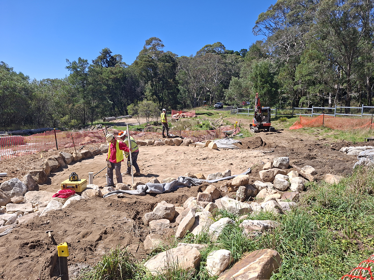 People working on a construction site, surrounded by rocks and orange safety fencing, there is an excavator in the distance and the construction site, is surrounded by bush landscape.