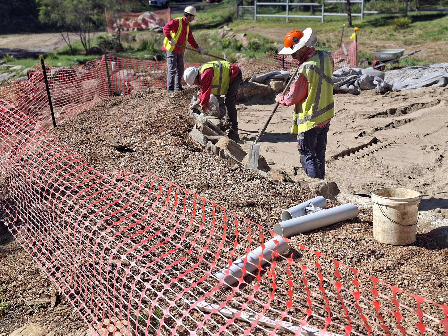 Three people working at a construction site, shovelling rocks, the construction site is surrounded by orange safety fencing.