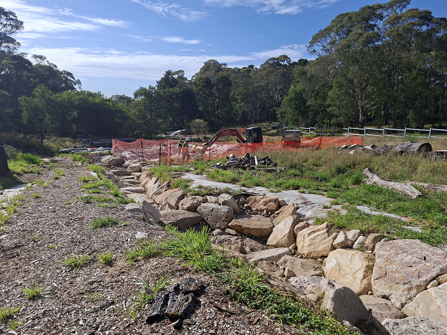 A construction site with people working and someone using an excavator, there is a long path of large rocks, and a bush landscape in the distance.