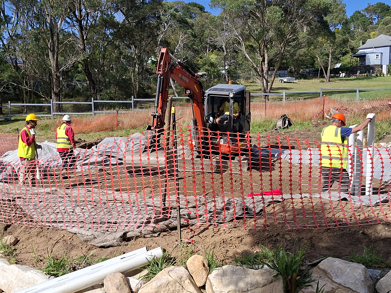 A group of people working at a construction site, one person is operating an orange excavator, there are large rocks and poly pipes on the ground.