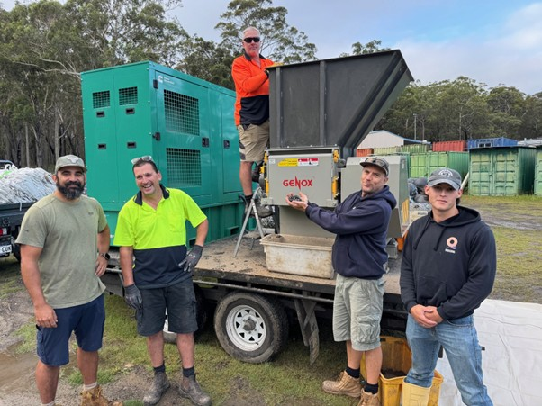 5 people looking at camera with four on the ground and one standing on back of trailer with machinery.