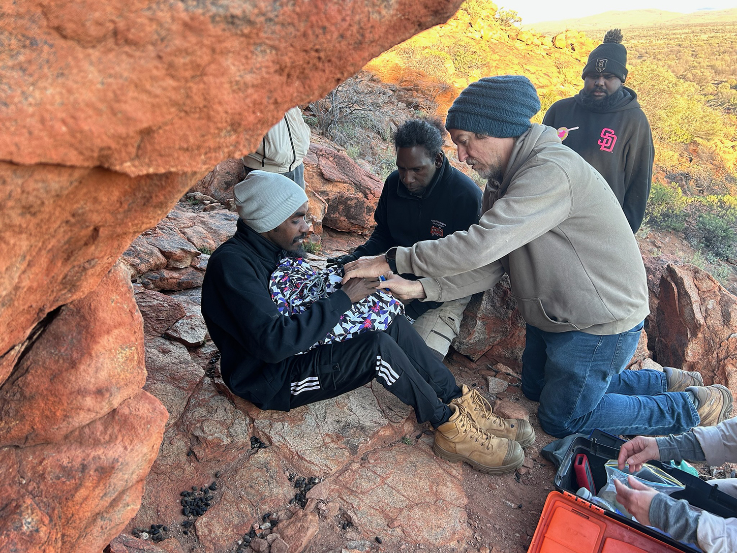 A group of people are sitting on red rocks, high above the ground, one person has an animal wrapped in a blanket.