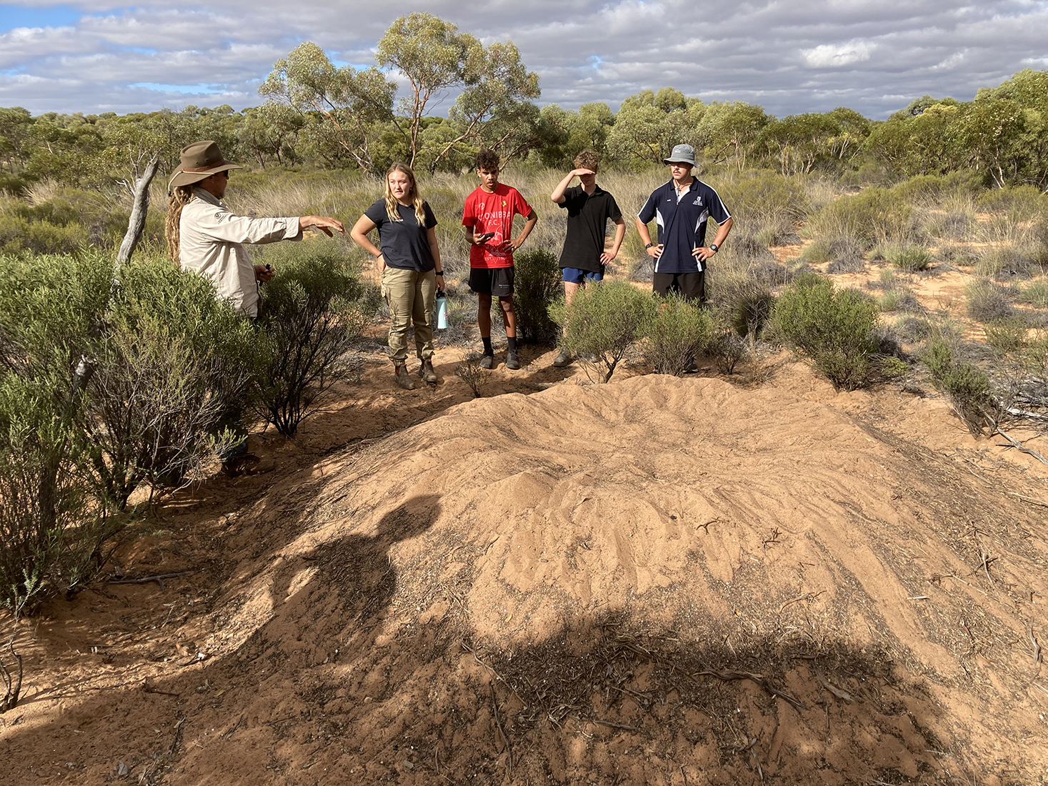 A group of people are standing around a dirt mound; there is a bush landscape behind them.