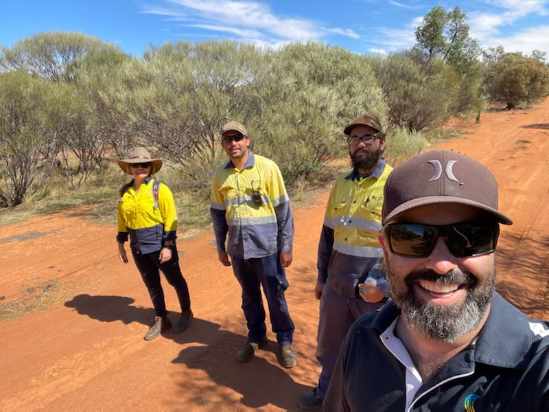 Group of four people in hi-vis facing the selfie camera standing on a red dirt track with bush in the background.