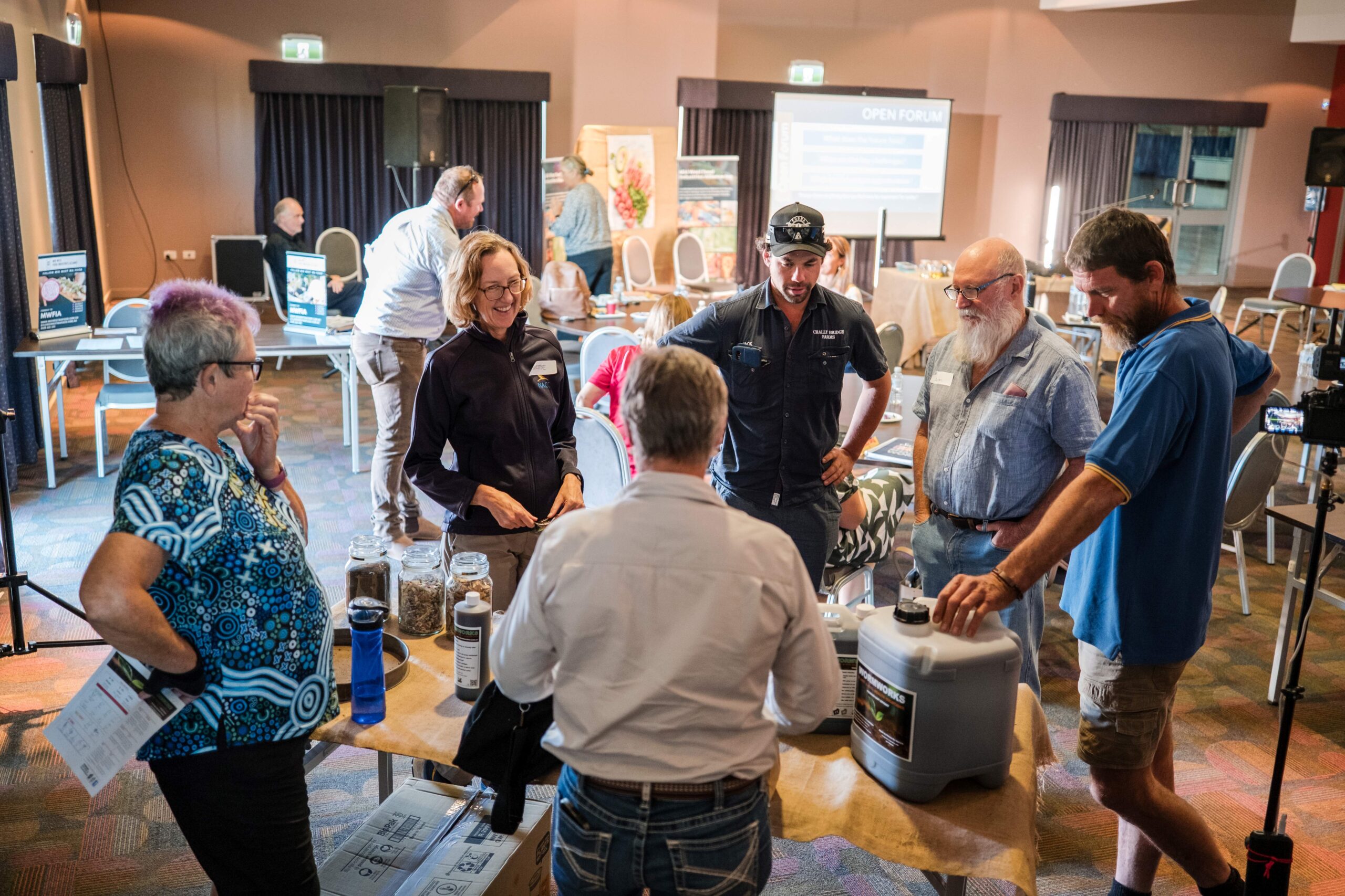 People standing grouped around a table in a room with other people and a screen in the background.