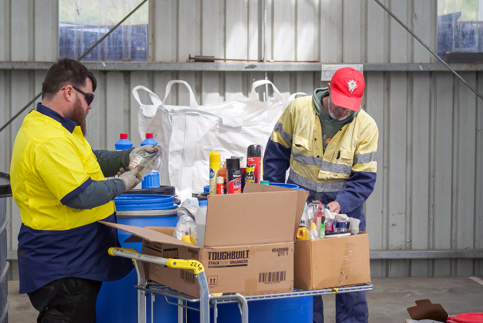 Two people standing in a shed sorting through boxes of hazardous household waste