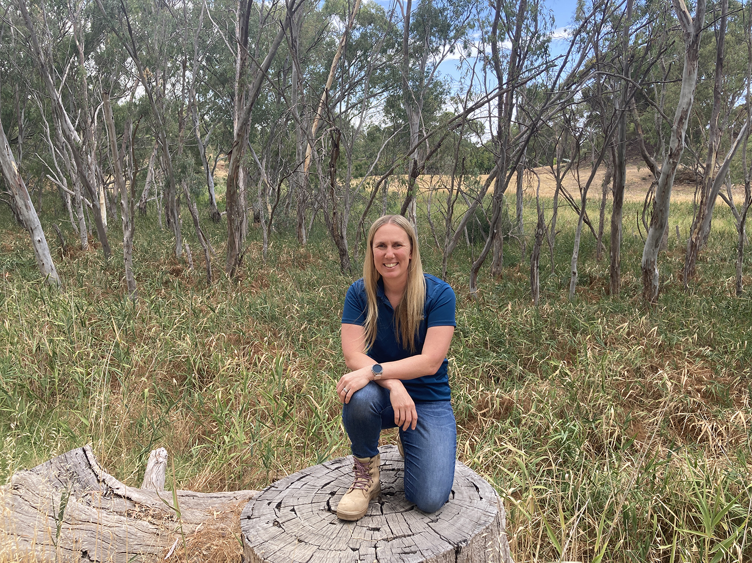 A person is kneeling on a tree stump, behind the person is a bush landscape.