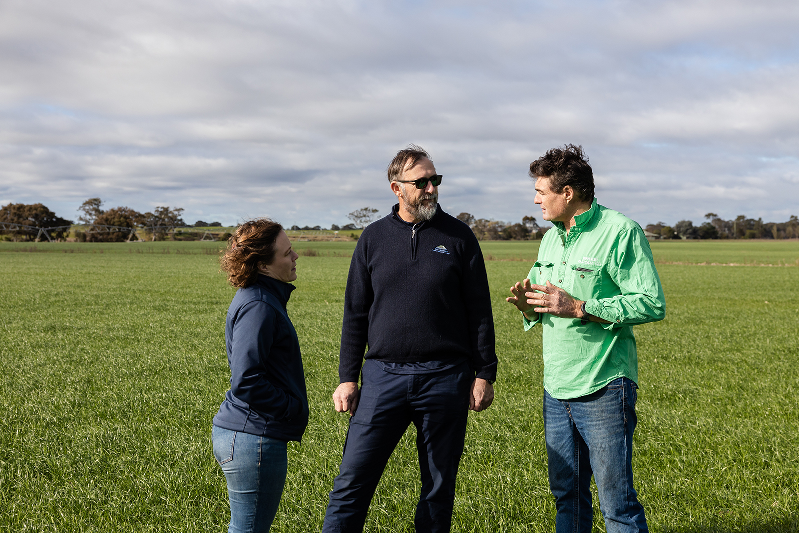 Three people standing in large grass covered farmland looking at each other with trees in the background.