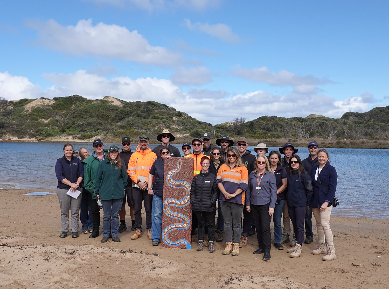 Large group of people standing facing the camera holding a piece of aboriginal artwork, standing on sandy bank of wide river with vegetated sand dune bank on other side.