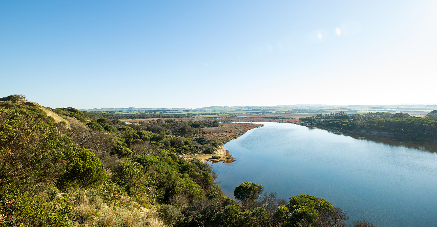 Landscape image looking down the length of a river with vegetated bushy banks in the foreground, reaching out to flat land and bare banks.