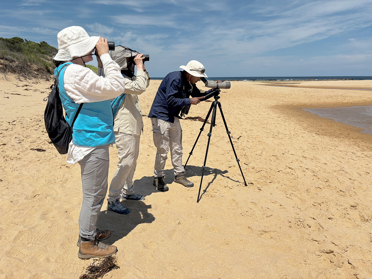 Three people standing on sandy bank of river near mouth to the ocean looking through binoculars