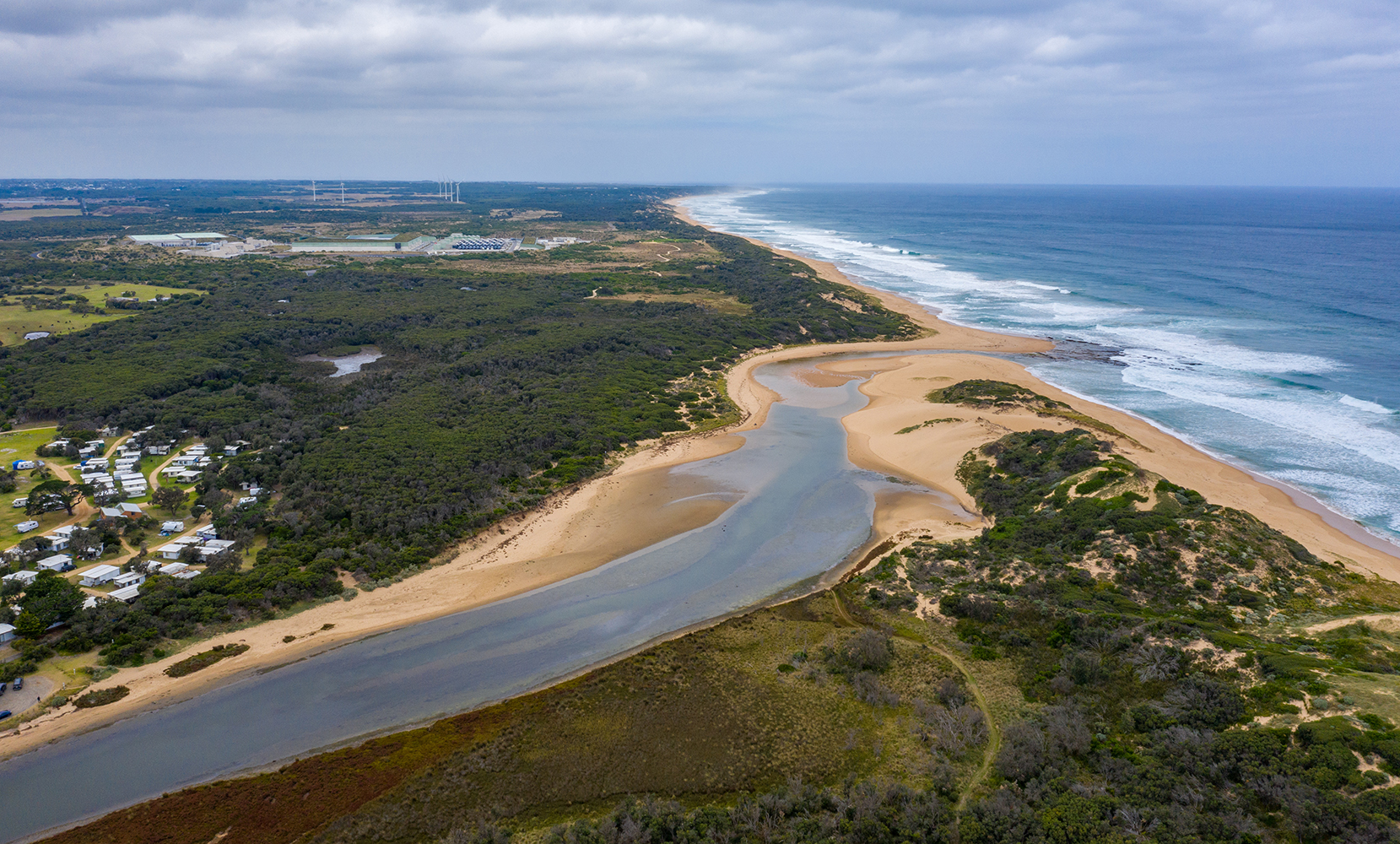 Aerial image of river and river mouth running out to ocean with sand banks, vegetation and a small town on the left.