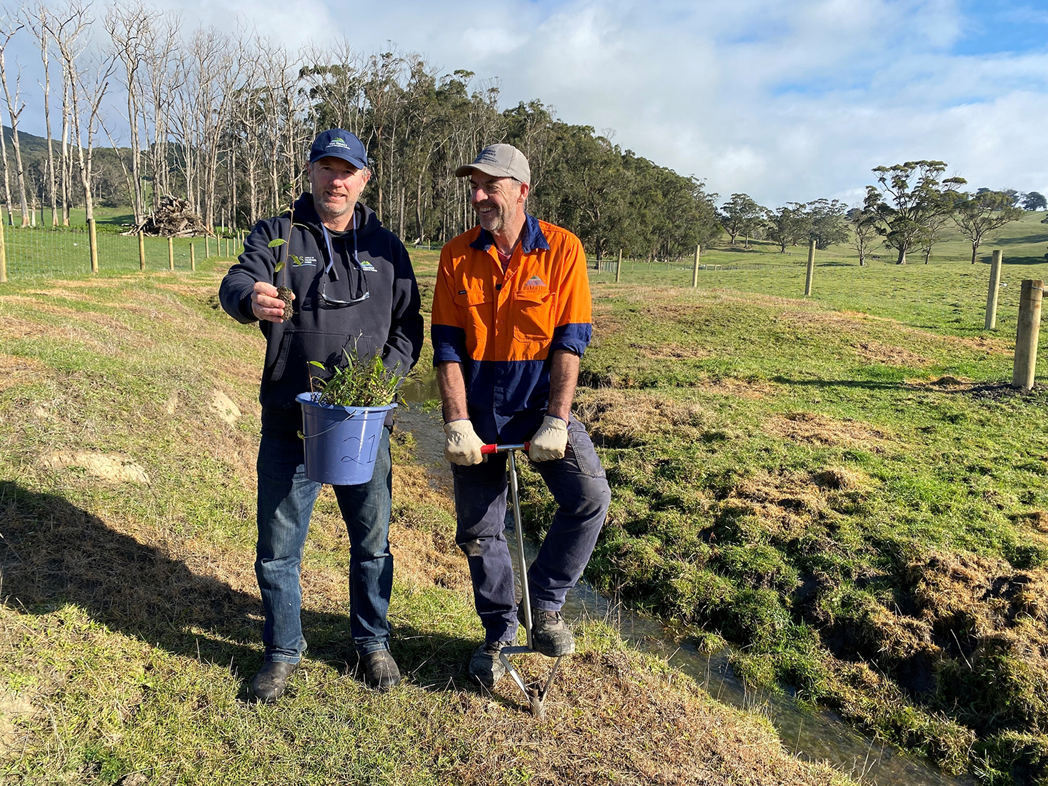 Two people with seedlings and planting equipment standing near a small stream near a fence with tall trees in the background.