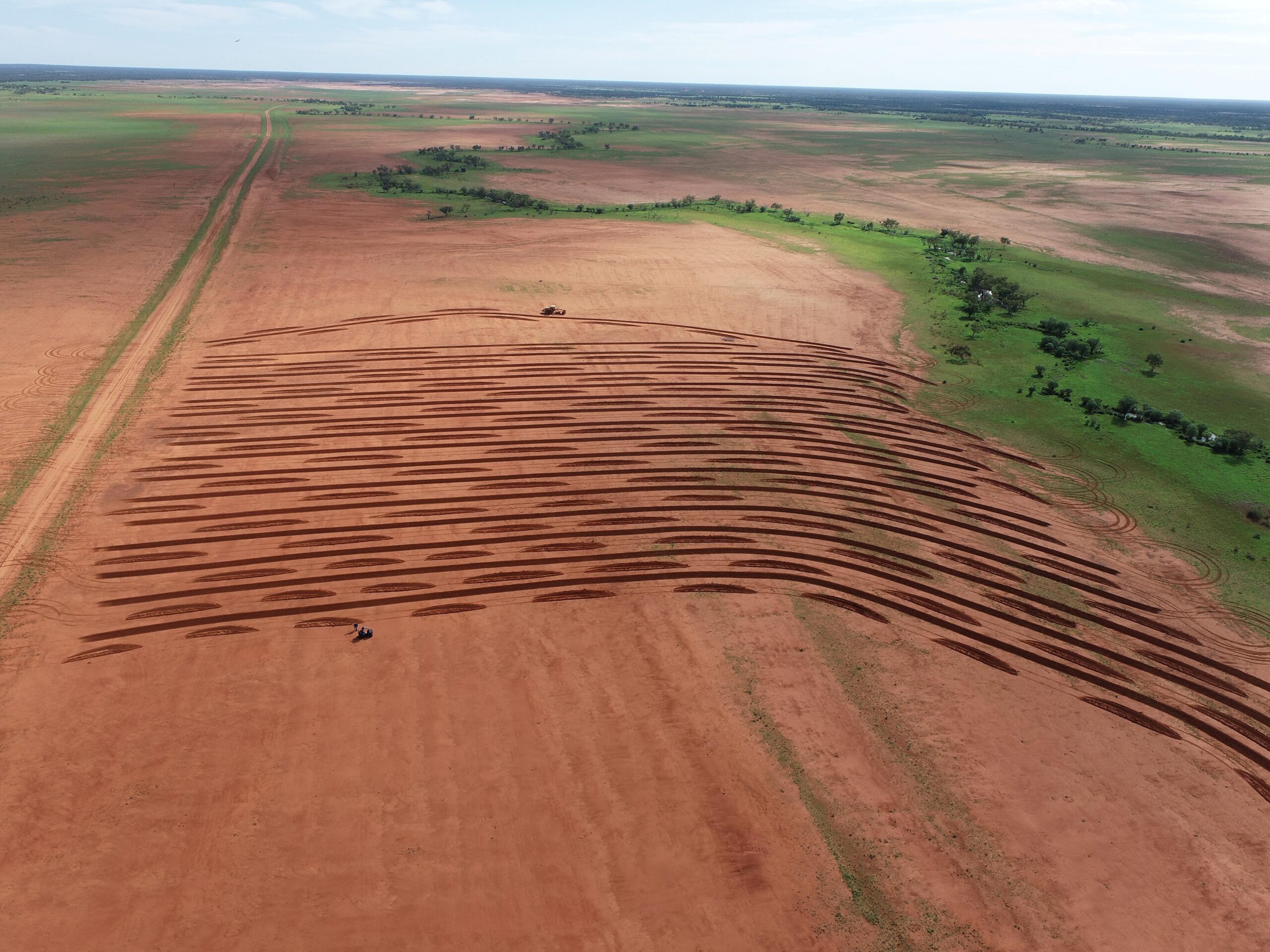 An aerial view of a large bare earth area with tractor ripping the ground in contour aligned brickwork furrows.