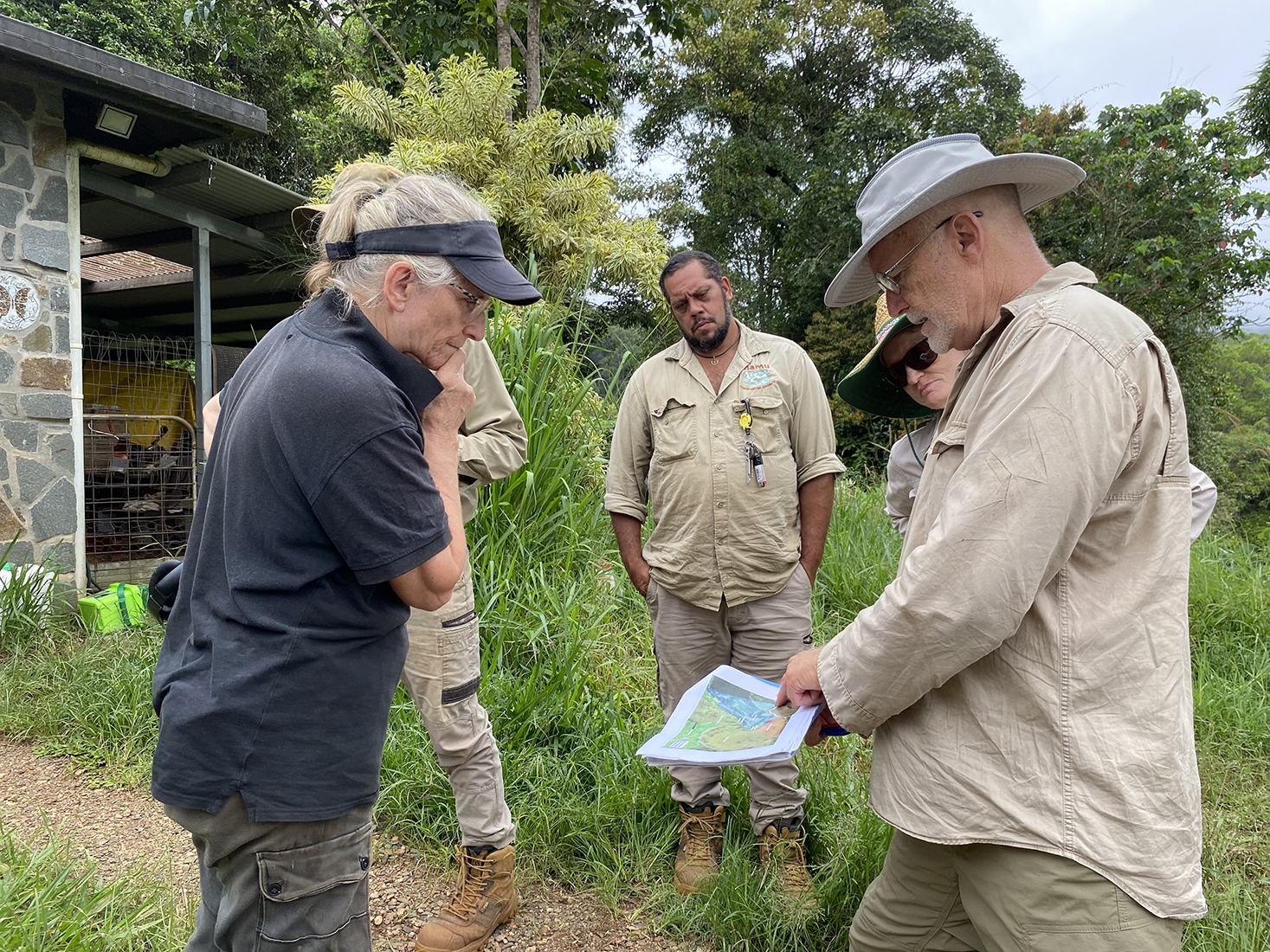 People are standing in a circle, looking at pieces of paper, with maps on them, they are surrounded by long, green grass and a brick building is behind them.