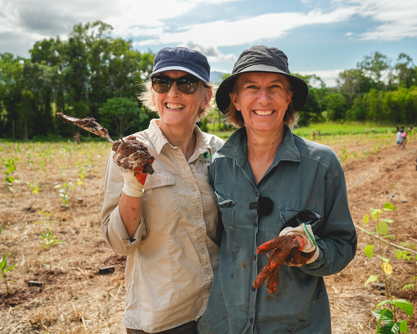 Two people are standing in a field, filled with tree saplings, they are holding muddy gloves and tools in their hands.