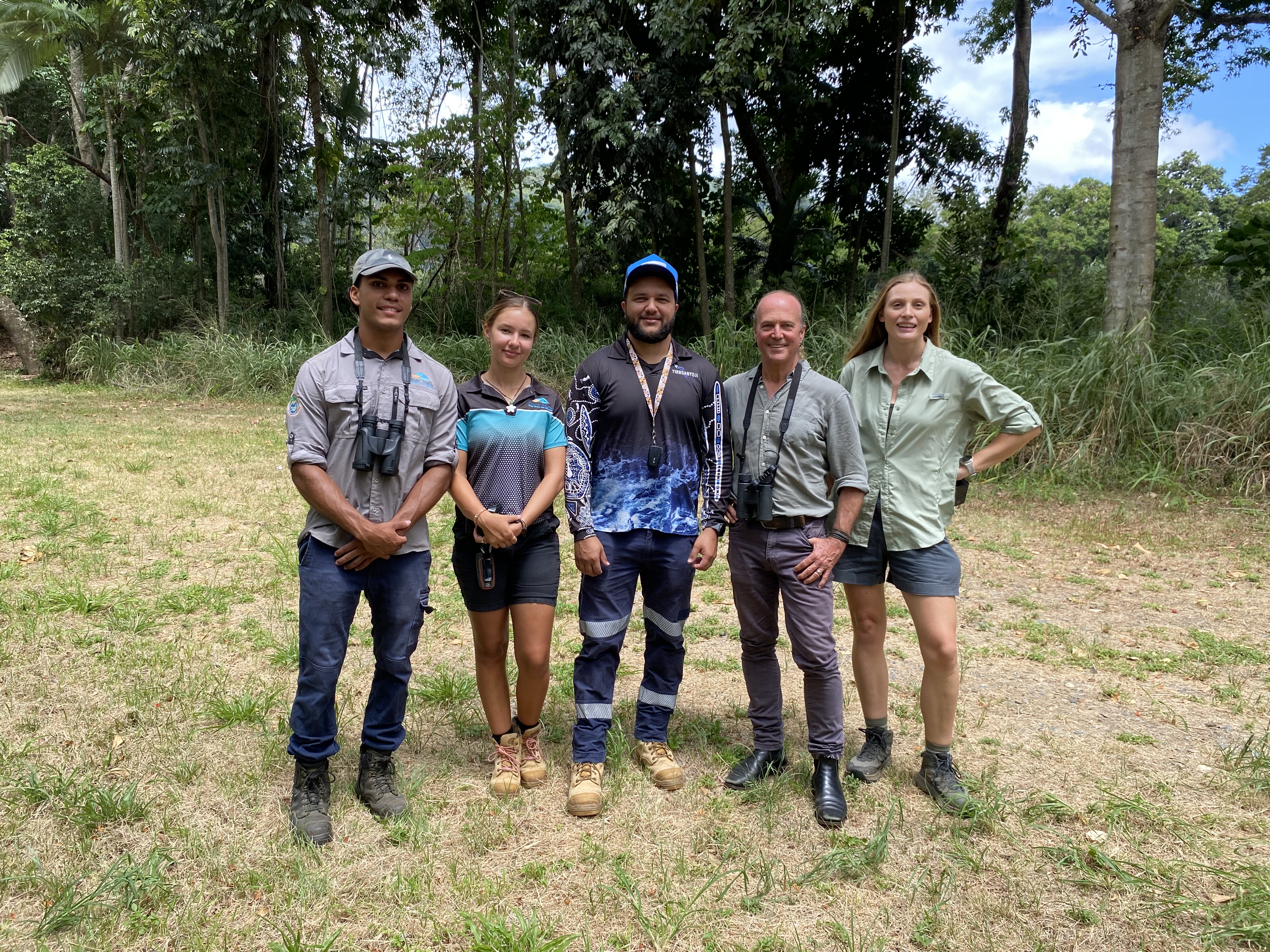 Five people some with binoculars looking at camera, smiling and standing on a clear area with dry grass and tufts of green with trees and vegetation in the background.