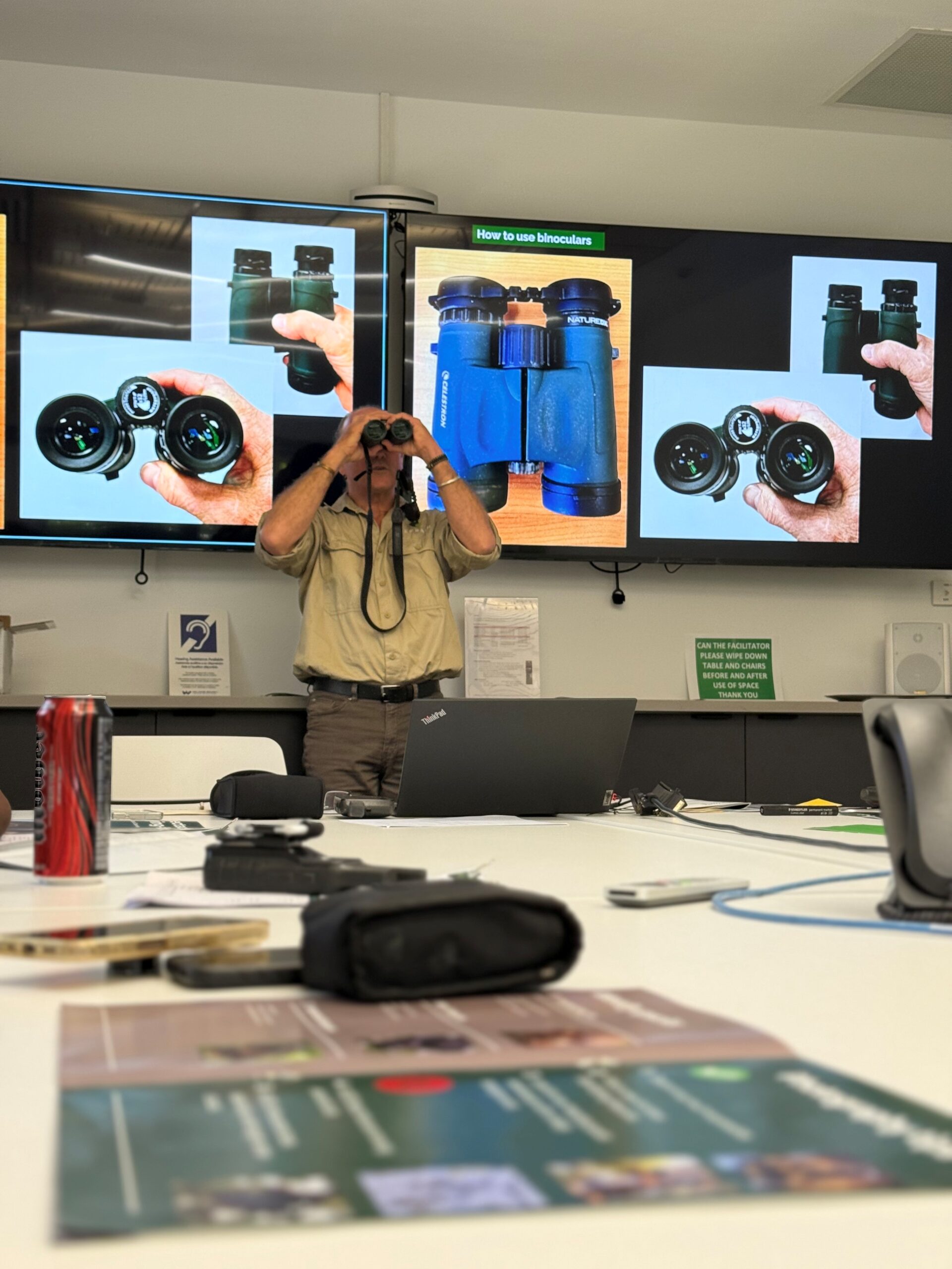 Person standing in room looking through binoculars with images of binoculars on screens behind him.