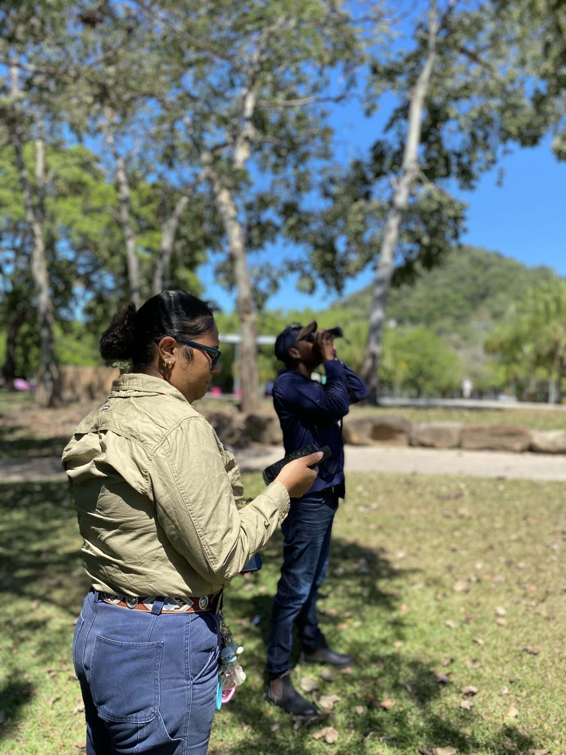 A person looking at a pair of binoculars in hand and another person standing looking through binoculars towards the sky, standing on a grassed area landscaped with rocks and trees in the background with a hill.
