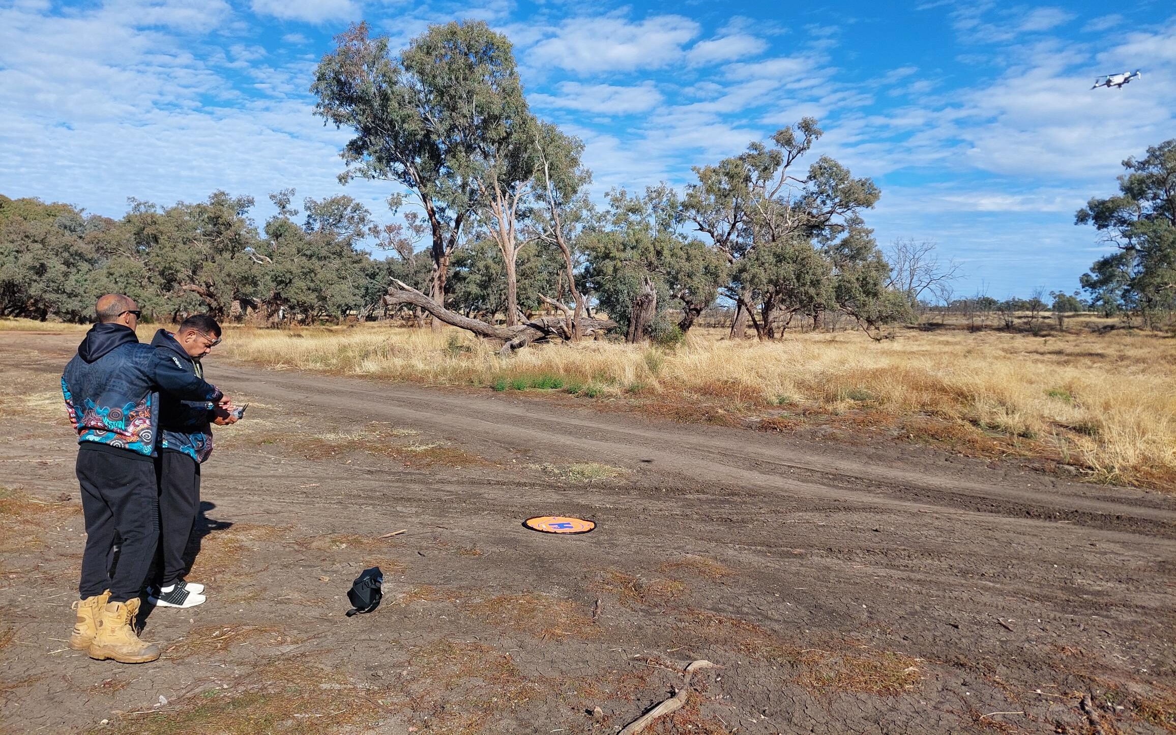 Two people learning to fly a drone with helipad in centre of image and drone top right in the bush.