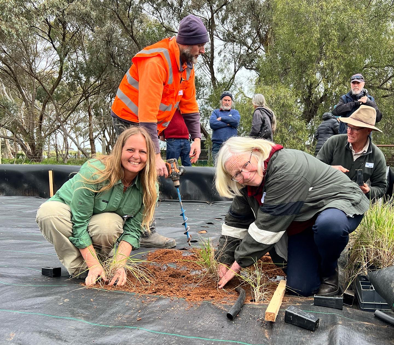Two people looking at the camera squatting planting vegetation in weed matting with more people behind, one with soil drill,