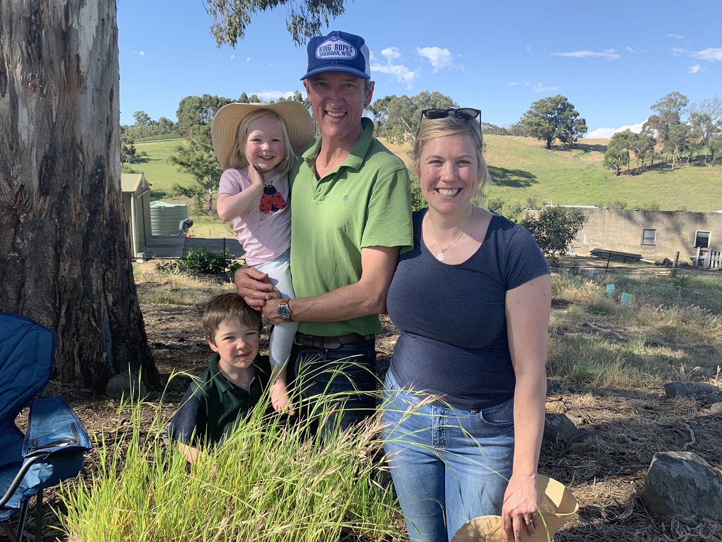 A family smiling looking at the camera standing by a tree with a tray of seedlings, buildings in the background green rolling hills dotted with trees.