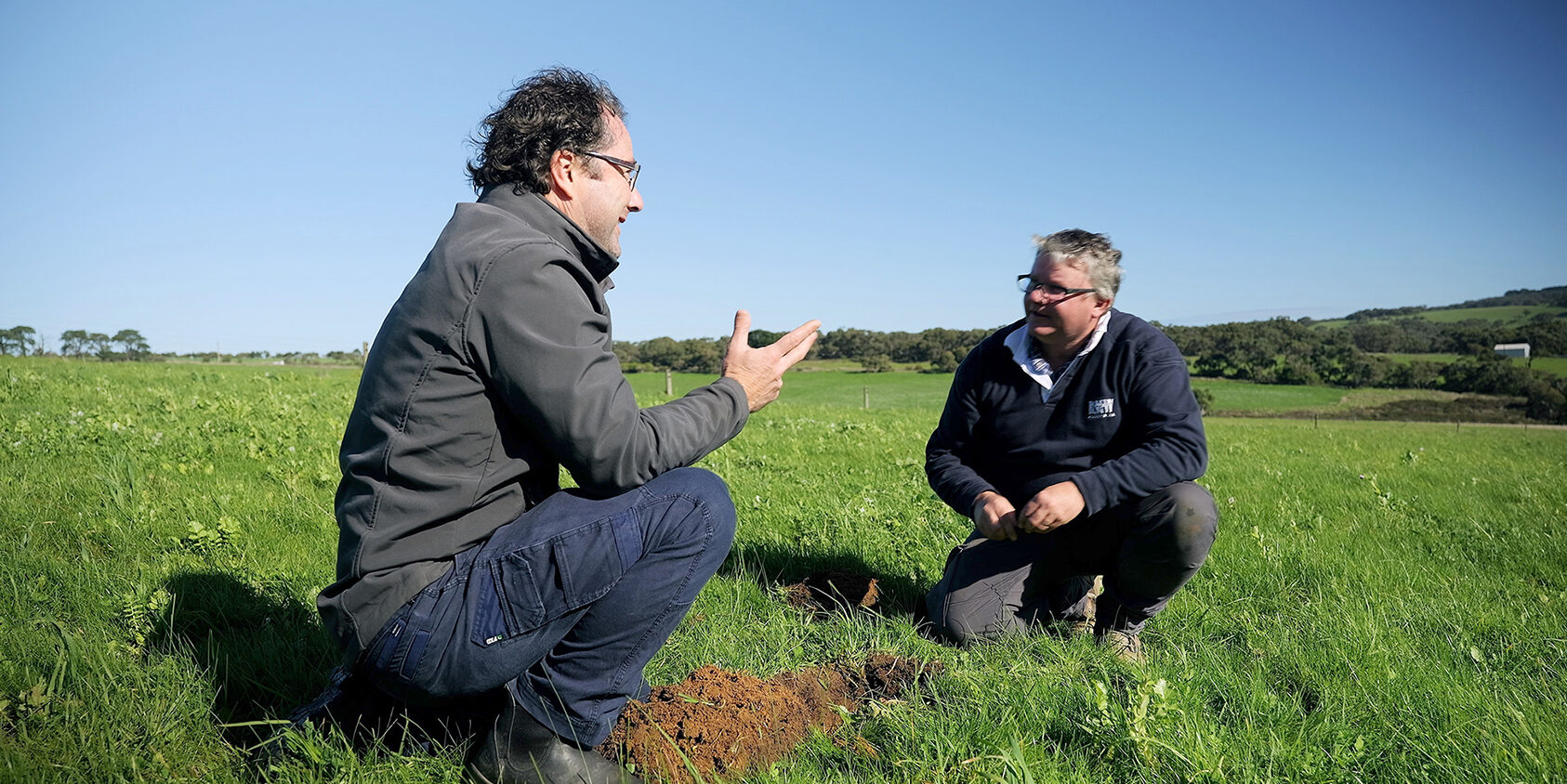 Two people looking at each other squatting on grass covered farm land with shrubs and trees in the distance.