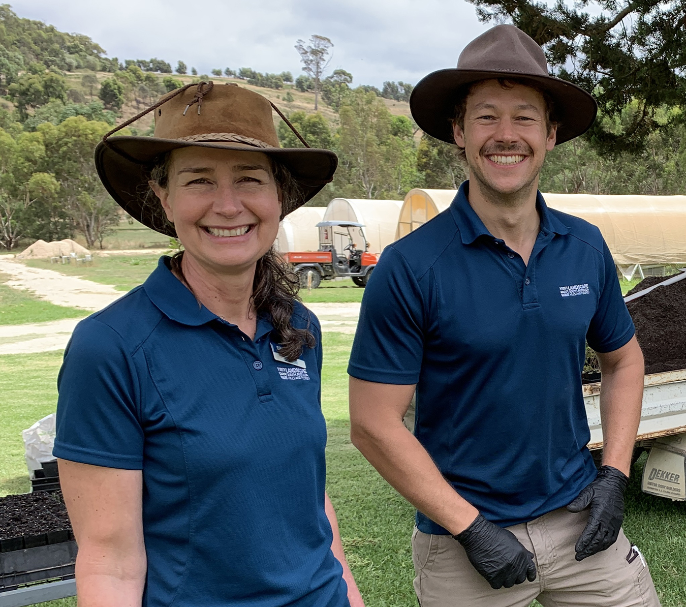 Two people smiling looking at the camera with hats on and one has gloves, standing near a vehicle with all terrain vehicle in the background and green houses