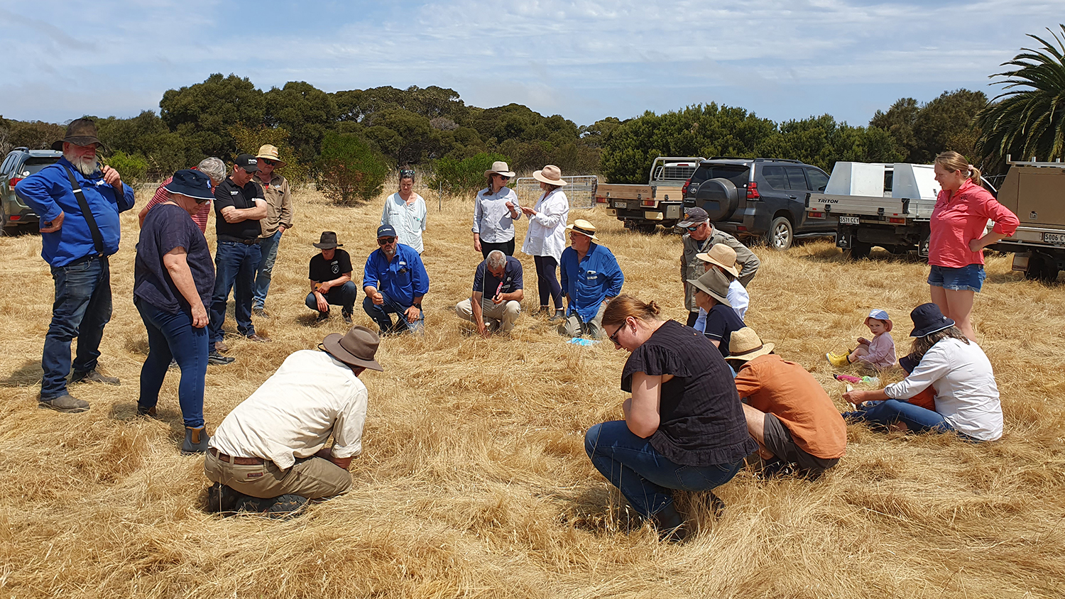 A group of people are in a dry, grassy field, there are four vehicles behind them and trees in the distance.