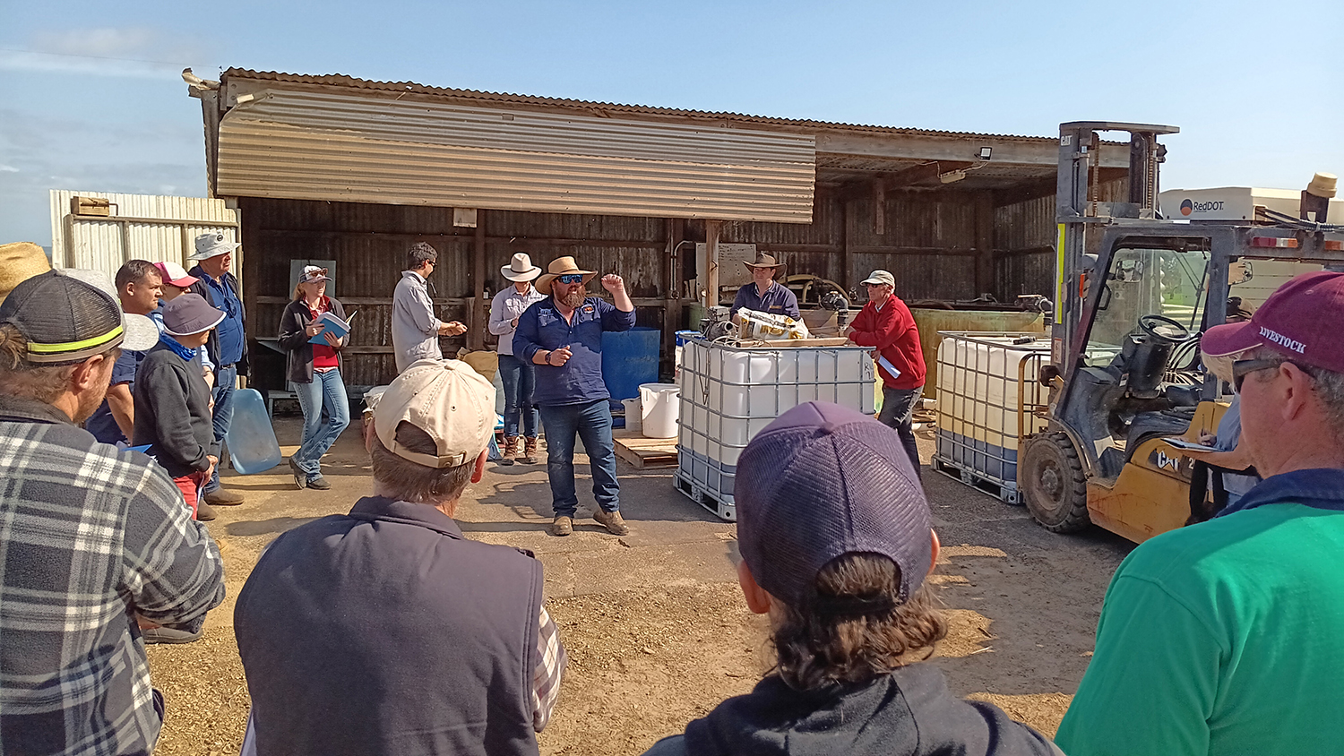 A group of people are watching a person talk, behind them is a shed, water tanks and a forklift.