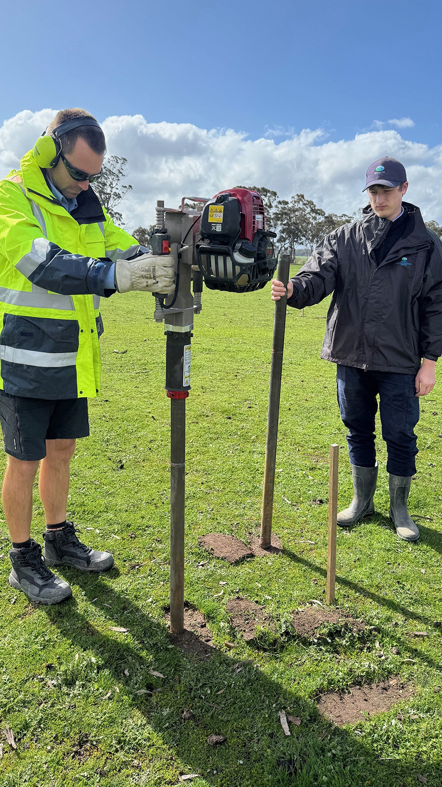 A person is drilling a pole into the ground, and another person is holding a pole.