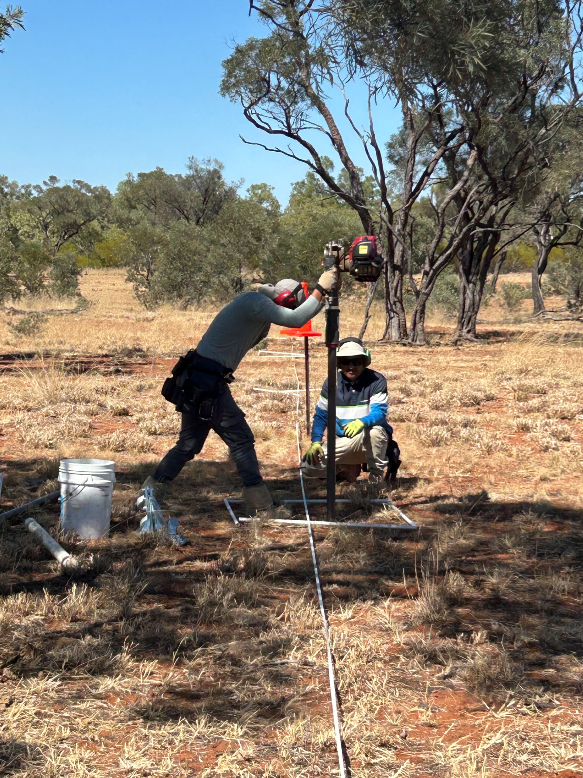 Two people are using large drill like machines, in a bush landscape.