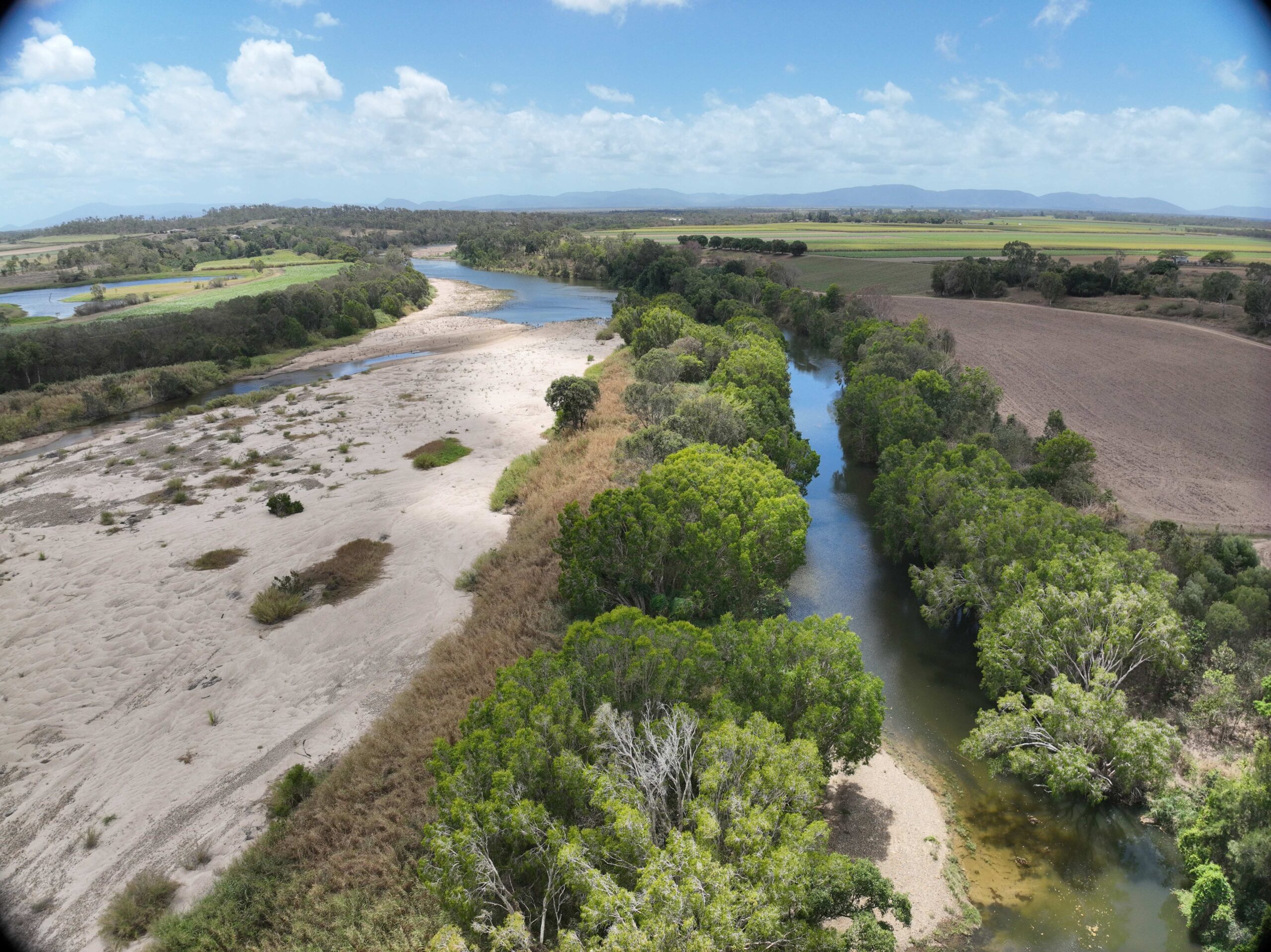 An aerial photo of a creek-line, flanked by green trees that give way to sandy soil.